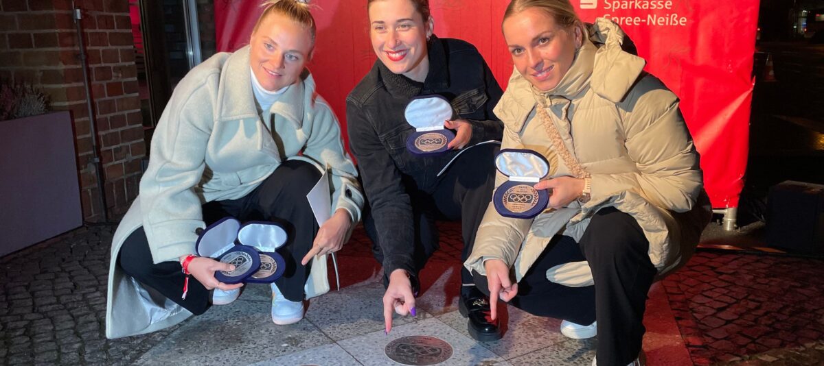 Pauline Grabosch, Lea Sophie Friedrich und Emma Hinze freuen sich über ihre Plaketten auf dem Weg des Ruhms in Cottbus und zeigen freudestrahlend mit dem Finger auf den Fußweg vor dem Cottbuser Rathaus im November 2024