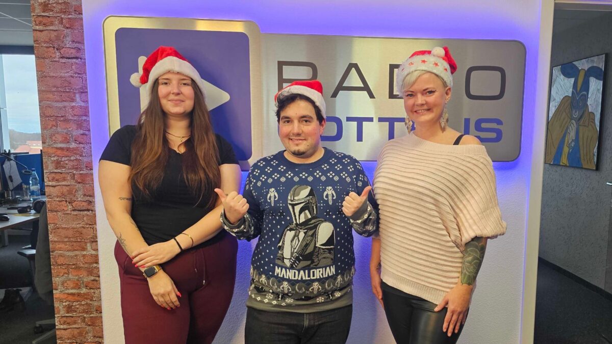 Nicole Liersch, Dena Nowak und Paulus Schinew vor dem Radio Cottbus Logo mit Weihnachtsmützen im Dezember 2024